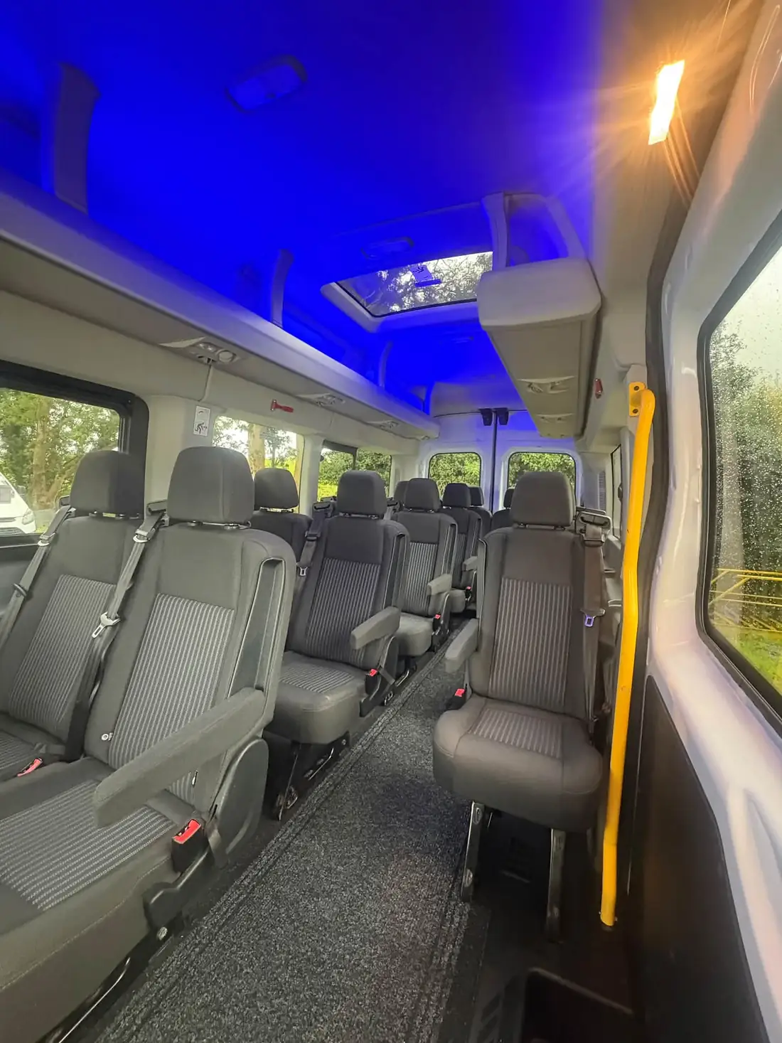 Inside the Abel Travel minibus: rows of grey reclining seats with armrests under blue ceiling lighting and an open roof window
